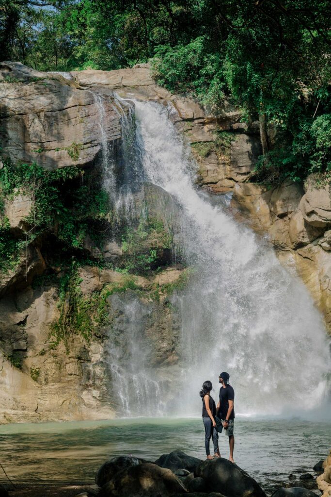 A boy and a girl dressed in black standing on a rocky terrain in front of Ellawala Falls, Sri Lanka