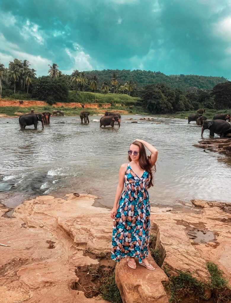 A girl wearing a blue frock on a rocky terrain in front of stream with few elephants