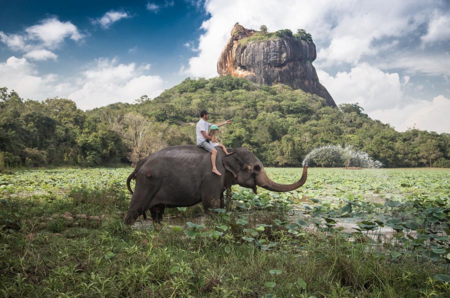 A father and a son on an elephant back, riding through a lake with the Sigiriya rock fortress far away